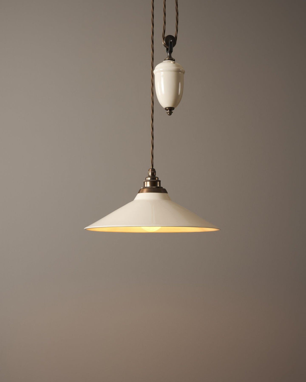 A rise and fall light with a small cream glazed ceramic shade and counterweight, an aged brass E27 lampholder and brown twisted fabric flex cord, the light is turned on. TEST