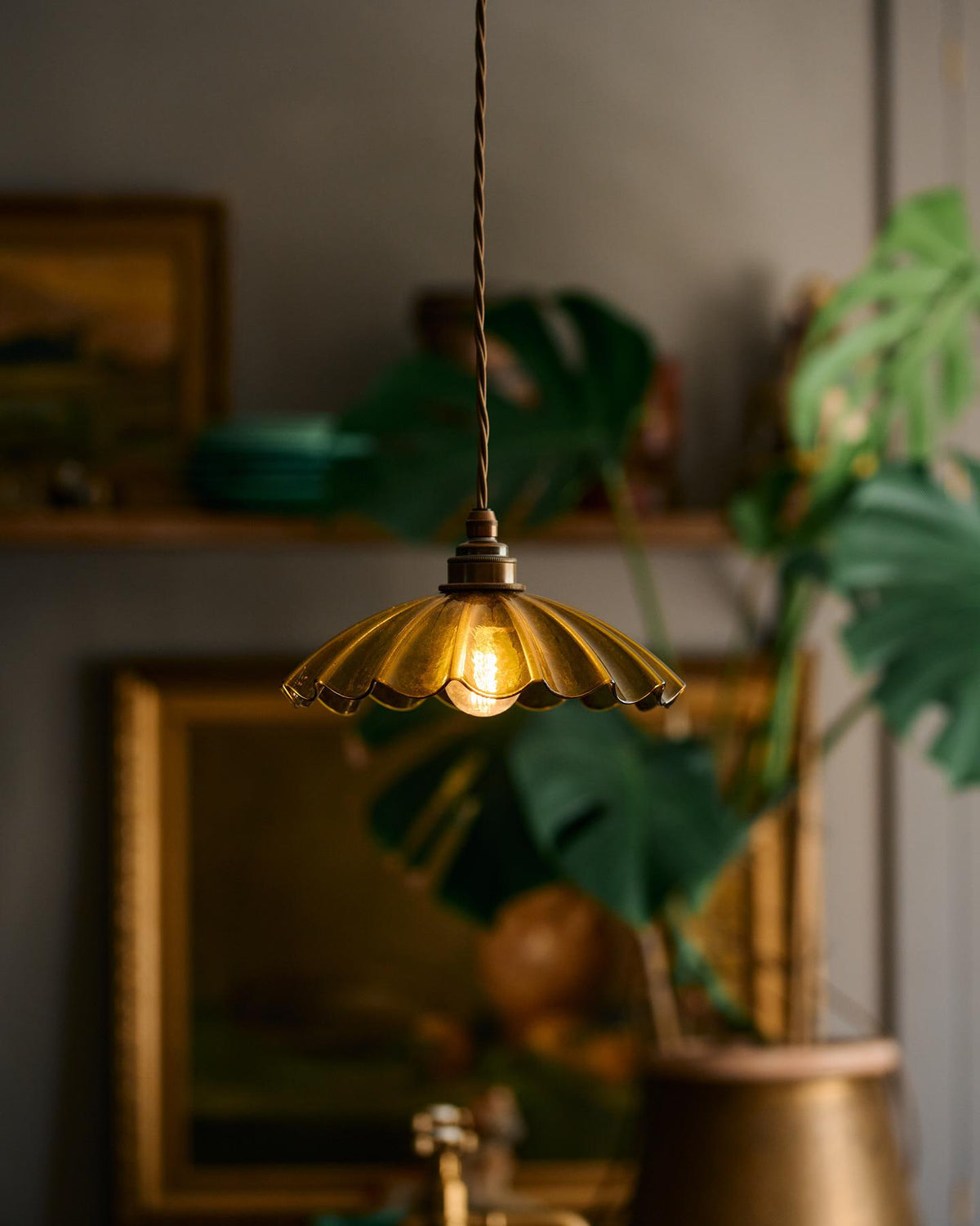 A close-up view of a small frilly chartreuse glass pendant light with brass fittings and brown twisted fabric flex cord. The light is on and shining through the tranlucent glass. TEST
