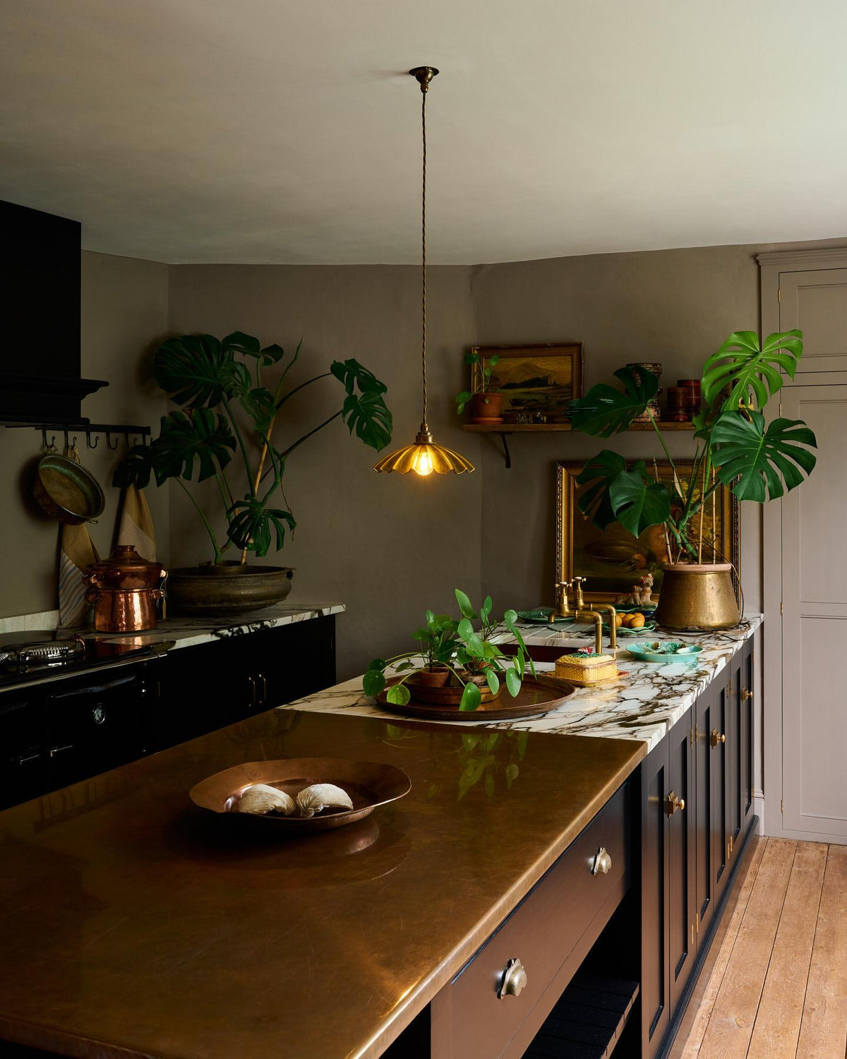 An oblique view of an atmospheric kitchen with black painted Shaker-style cabinetry, grey walls, wooden floorboards, vintage styling and large monstera plants. A small frilly chartreuse glass pendant light with brass fittings and brown twisted fabric flex cord is hanging above an aged copper worktop. TEST