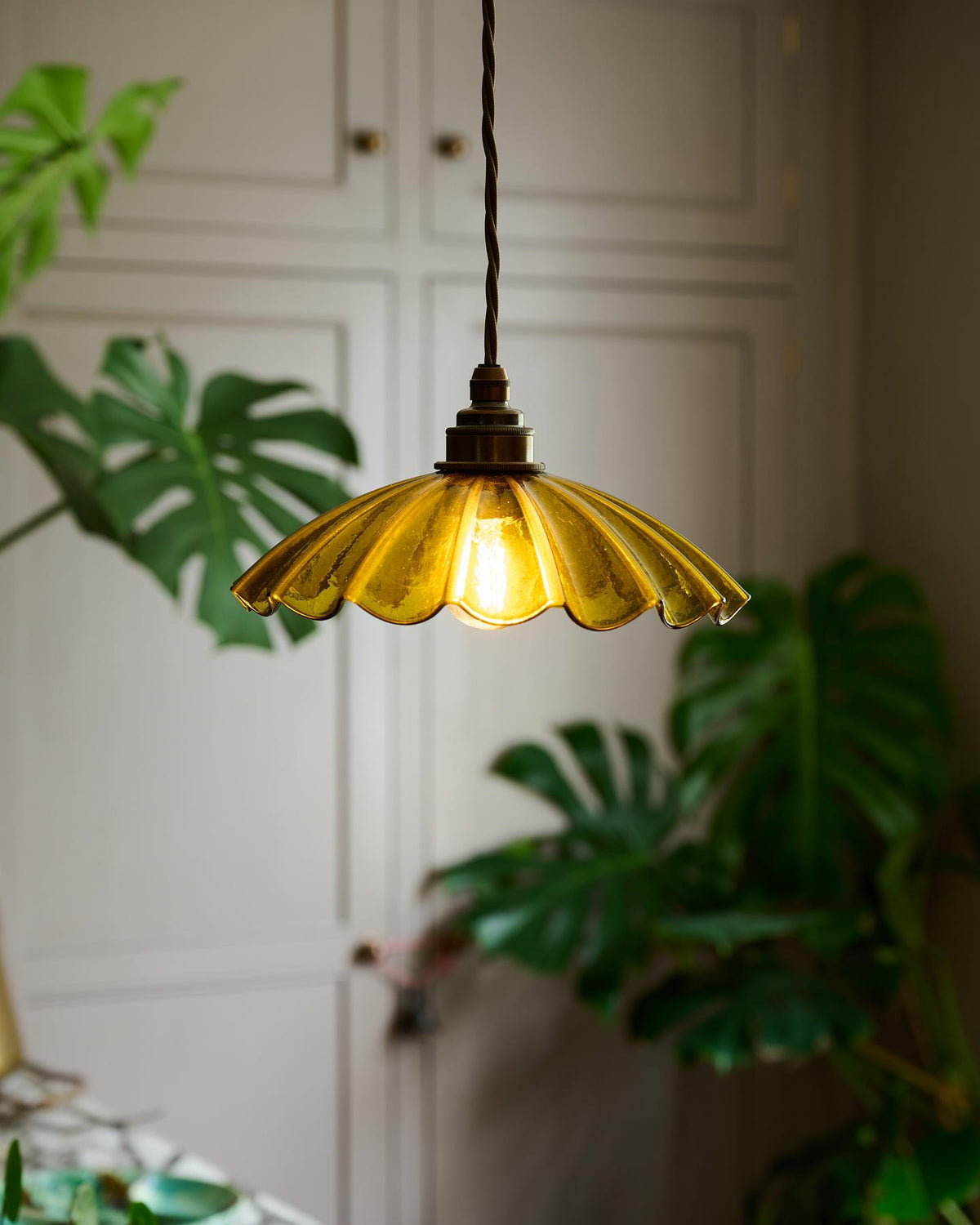 A small frilly chartreuse glass pendant light with brass fittings and brown twisted fabric flex cord. The light is on and shining through the tranlucent glass. TEST