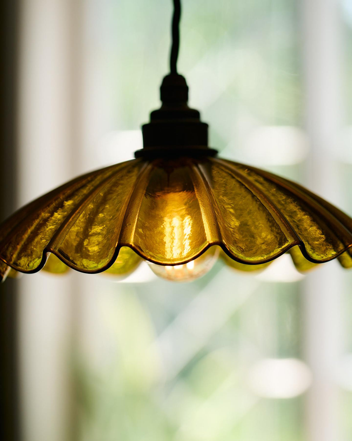 A close-up view of a small frilly chartreuse glass pendant light with brass fittings and brown twisted fabric flex cord. The light is on and shining through the tranlucent glass. TEST