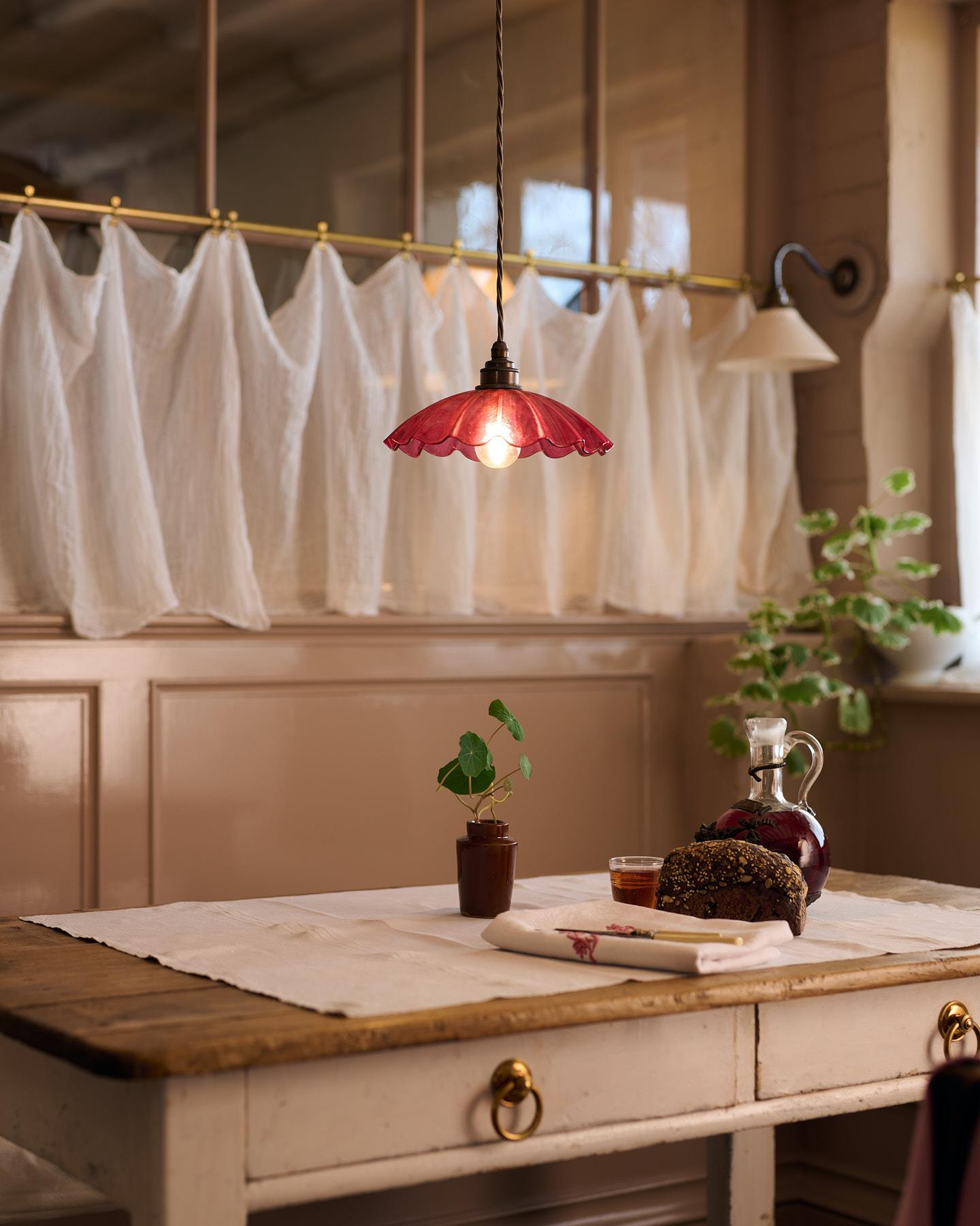 An oblique view of a small frilly cranberry glass pendant light with brass fittings and brown twisted fabric flex cord hanging above an antique pine table in front of a glazed partition with a lace curtain.