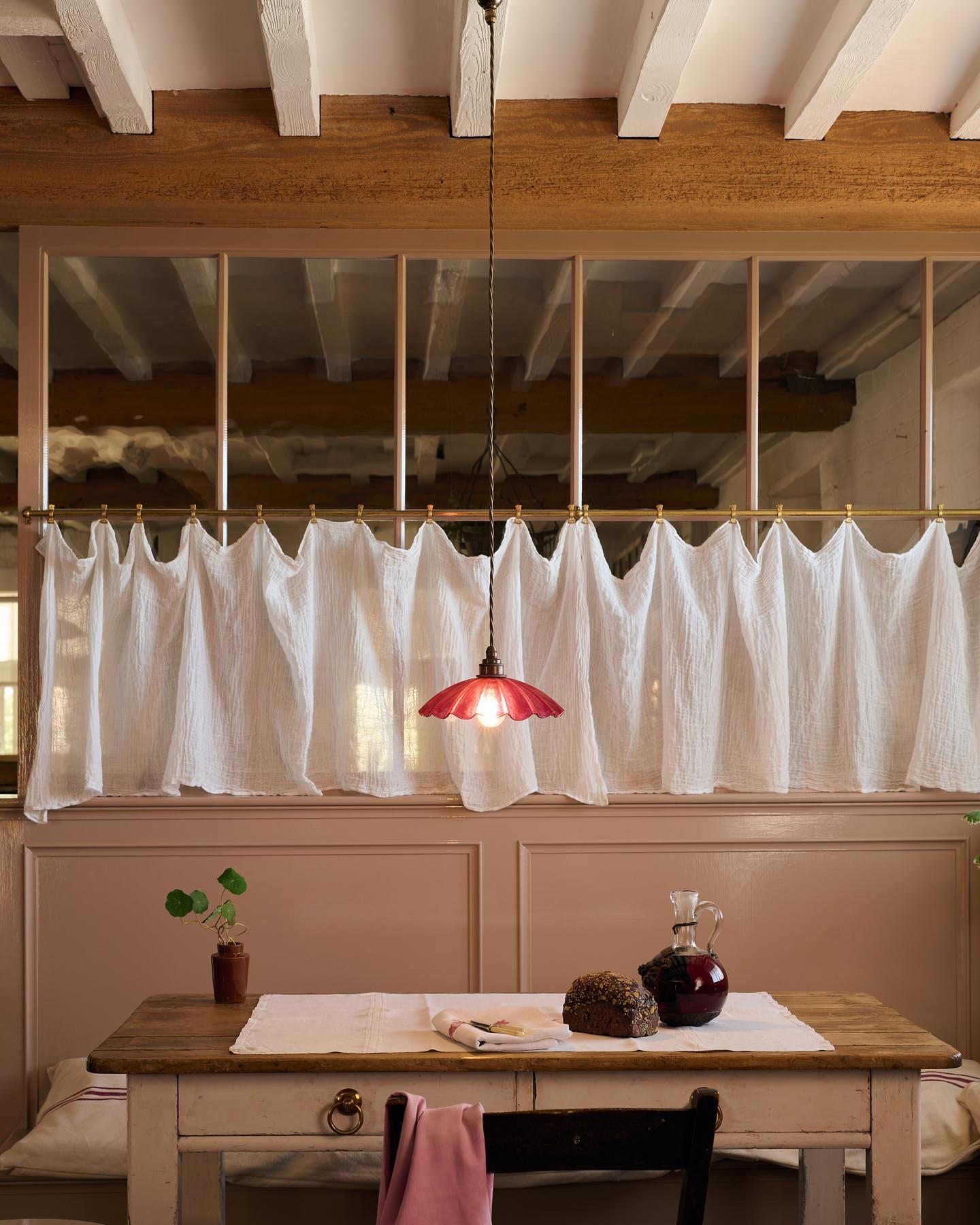 A straight-on view of a small frilly cranberry glass pendant light with brass fittings and brown twisted fabric flex cord hanging above an antique pine table in front of a glazed partition with a lace curtain.