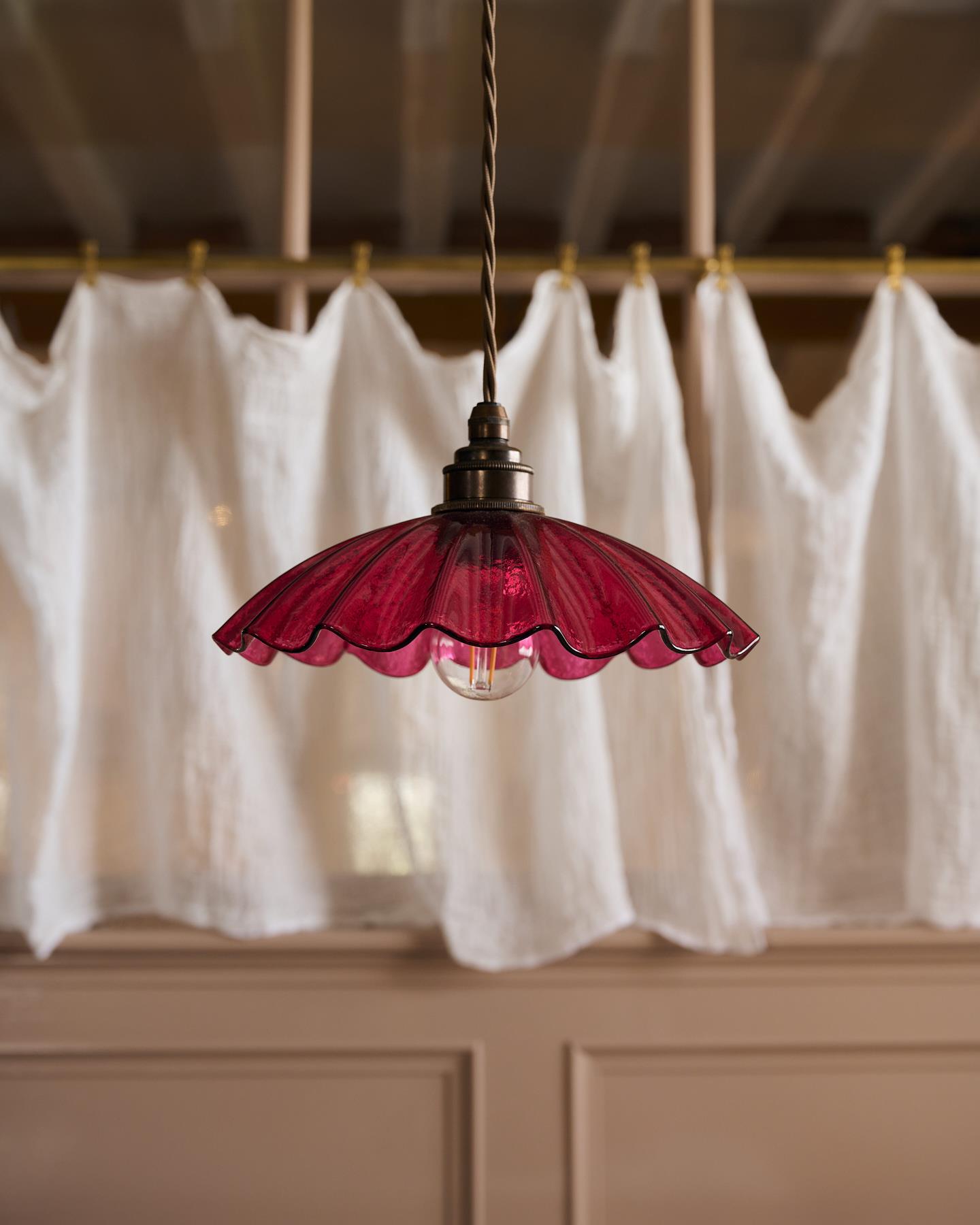 A close-up view of a small frilly cranberry glass pendant light with brass fittings and brown twisted fabric flex cord. The light is off.