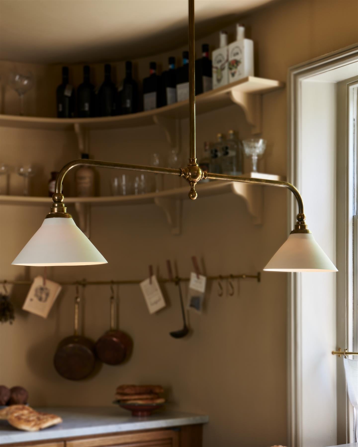An oblique view of a mid-century-style kitchen with oak cabinetry, buttery yellow walls and a Victorian-inspired chandelier light with brass arms and small unglazed porcelain shades over a prep table with a Carrara marble worktop.