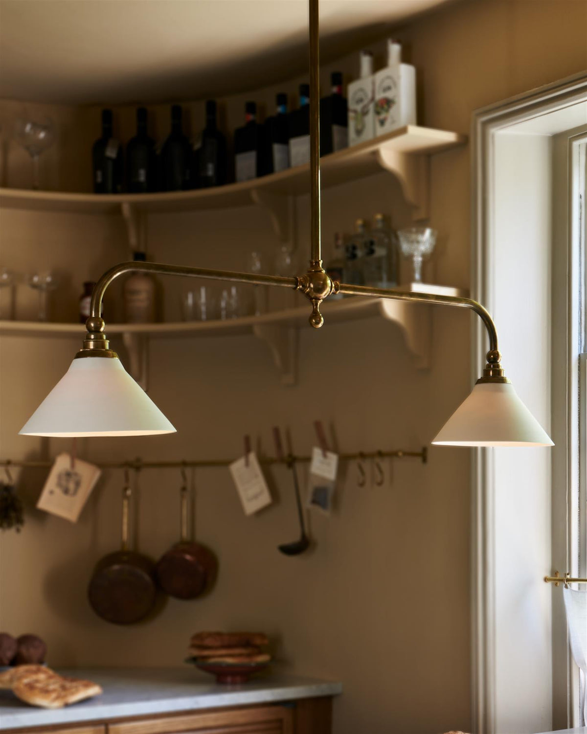 An oblique view of a mid-century-style kitchen with oak cabinetry, buttery yellow walls and a Victorian-inspired chandelier light with brass arms and small unglazed porcelain shades over a prep table with a Carrara marble worktop. TEST