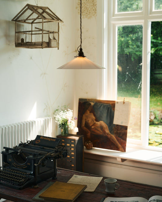 An oblique view of a classic pendant light with a milk glass light shade, brass fittings and a cream twisted fabric flex cord, hanging above a typewriter and scraps of paper by an old oil painting and a large window.