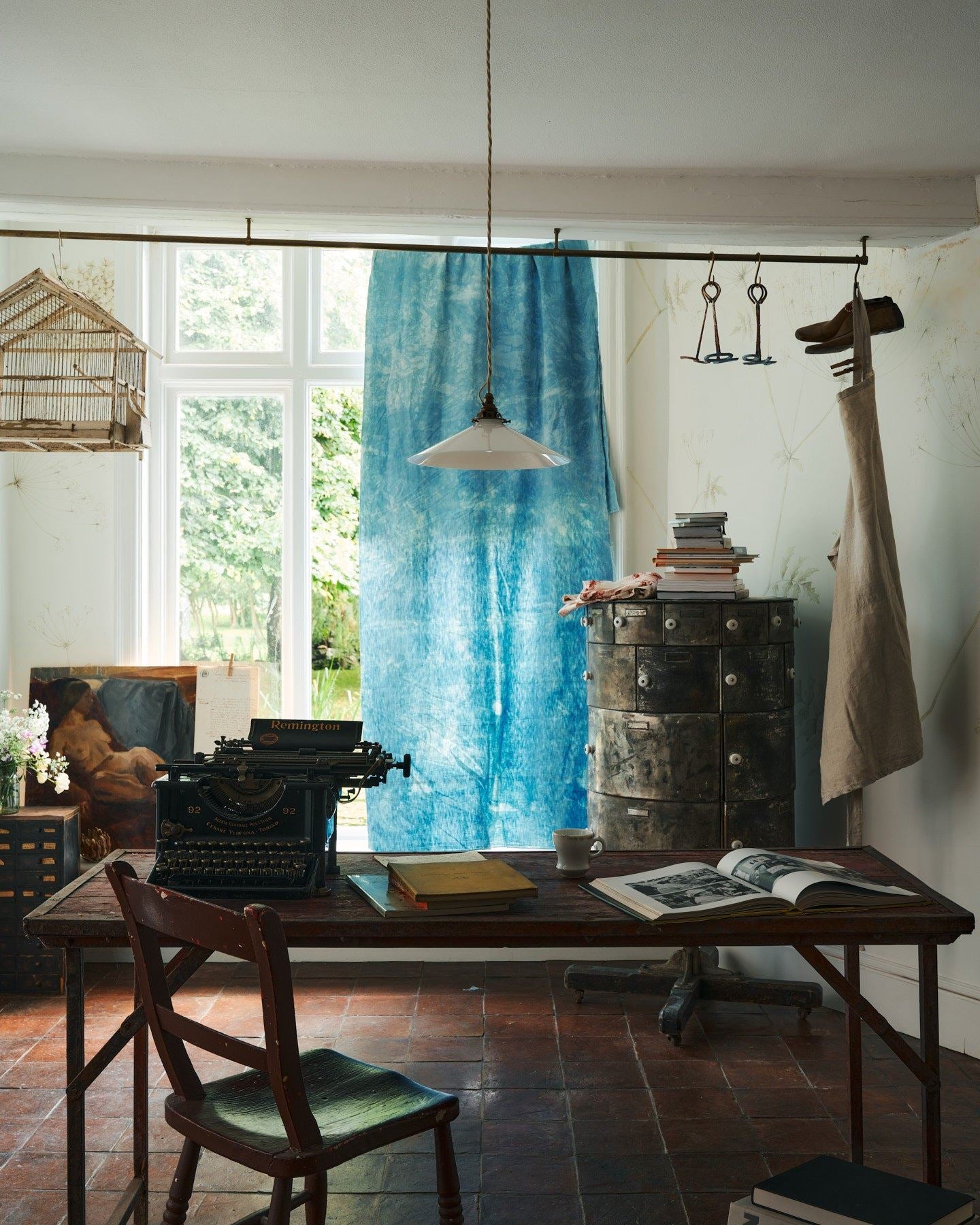 A straight-on view of a classic pendant light with a milk glass light shade, brass fittings and a cream twisted fabric flex cord, hanging above a typewriter and scraps of paper.