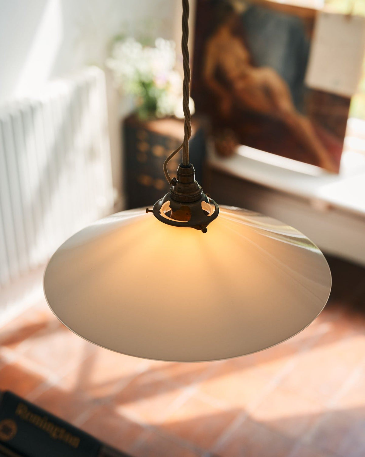 A close-up oblique view of a classic pendant light with a milk glass light shade, brass fittings and a cream twisted fabric flex cord. The light is on, showing the warm red glow of the translucent surface. TEST
