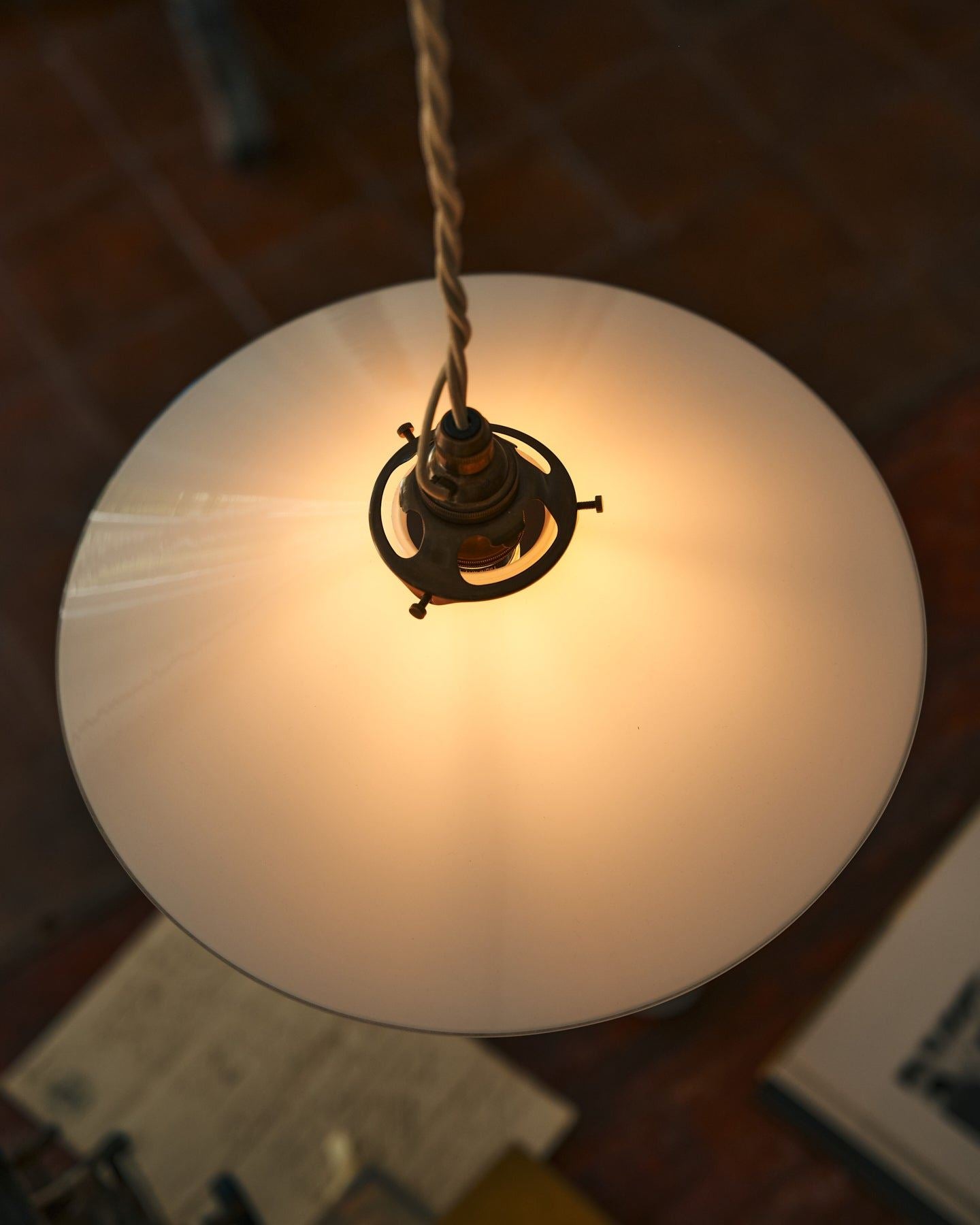 A close-up overhead view of a classic pendant light with a milk glass light shade, brass fittings and a cream twisted fabric flex cord. The light is on, showing the warm red glow of the translucent surface.