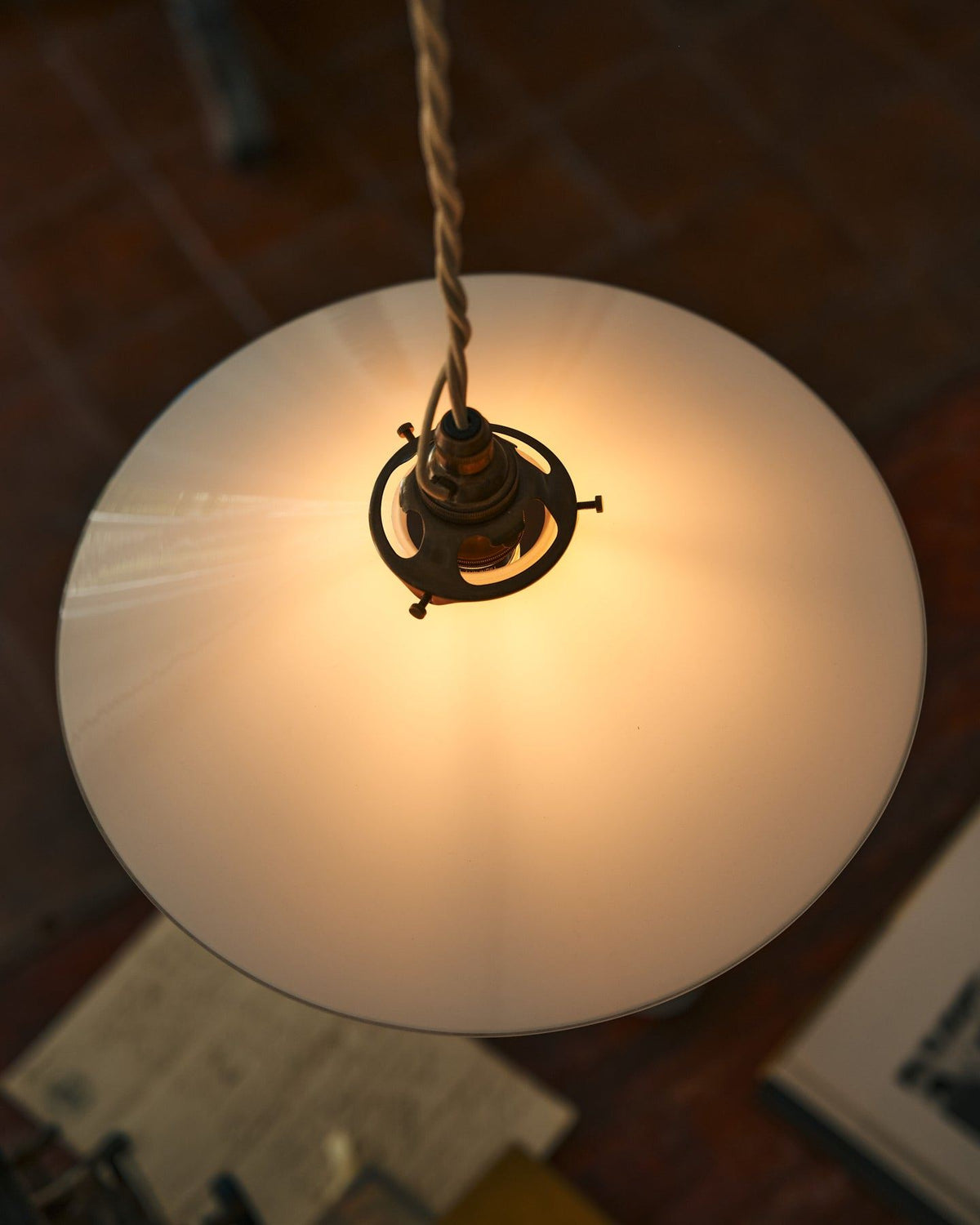 A close-up overhead view of a classic pendant light with a milk glass light shade, brass fittings and a cream twisted fabric flex cord. The light is on, showing the warm red glow of the translucent surface. TEST