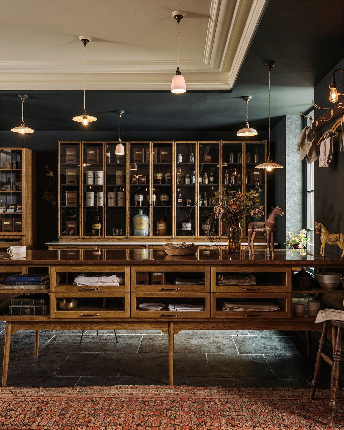 A straight-on view of a mid-century-style kitchen with oak cabinetry, dark painted walls and an assortment of unglazed porcelain pendant lights hanging at various heights. TEST