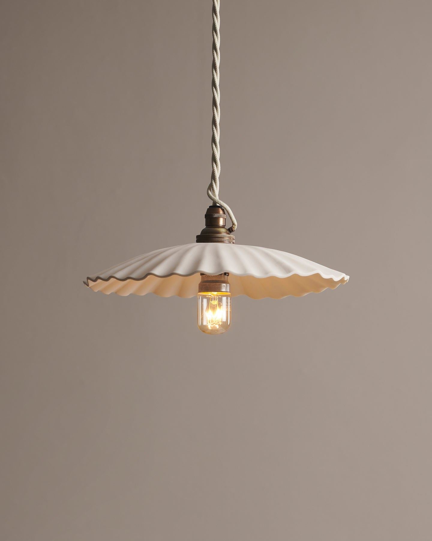 A large narrow pleat porcelain pendant light with an unglazed finish, an aged brass B22 lampholder and a white twisted fabric flex cord.