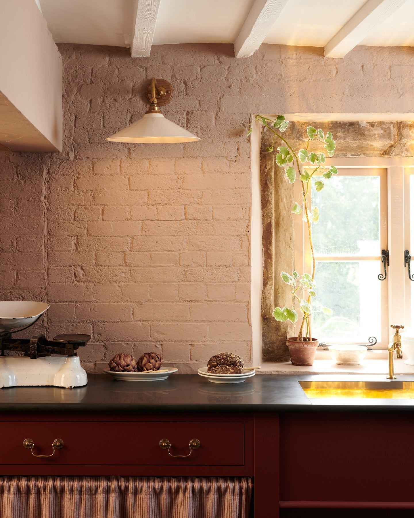 A straight-on view of a large Victorian-inspired wall light with a brass arm and a cream glazed ceramic shade fixed to a pink painted brick wall above a slate worktop and red painted cabinetry.