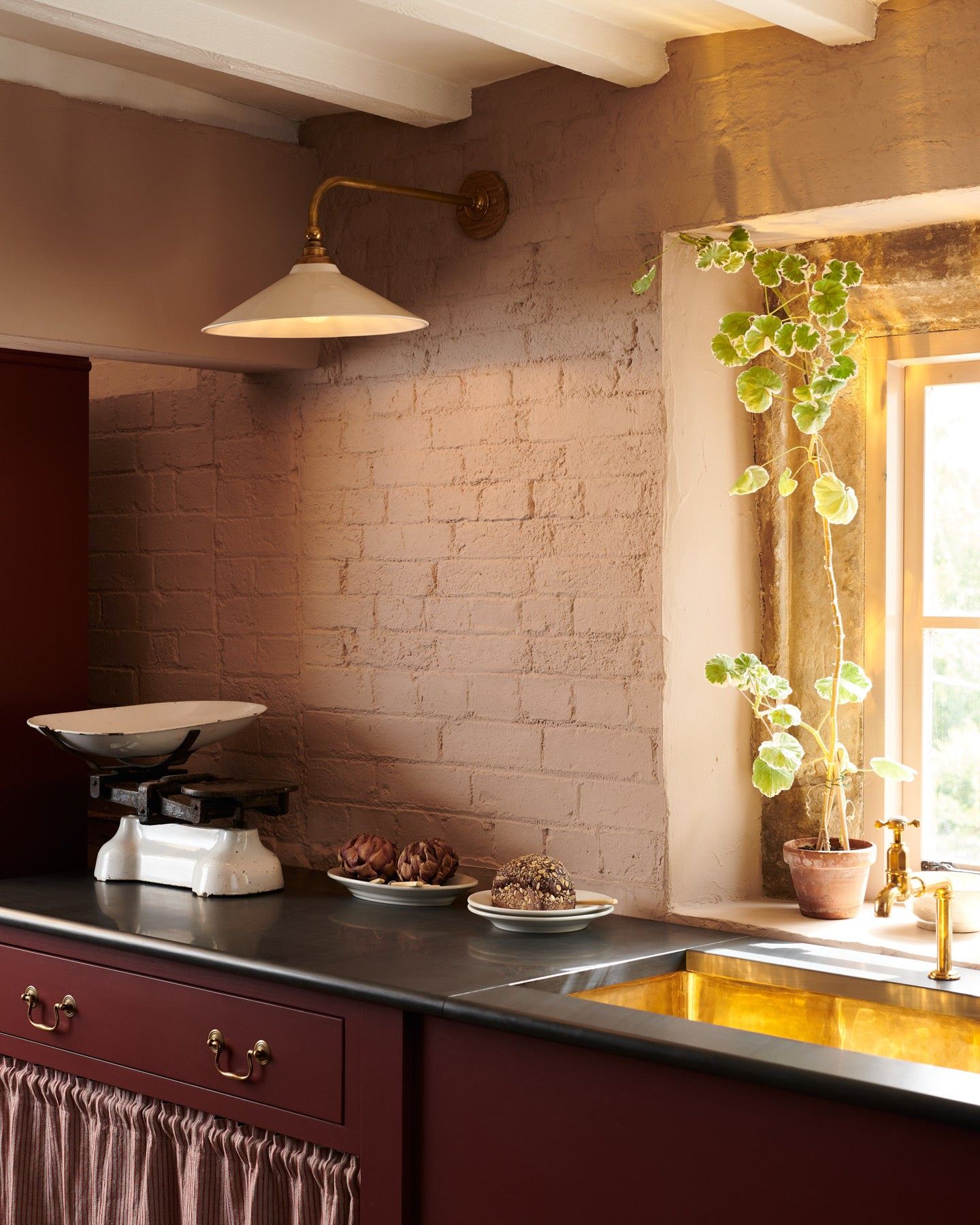 An oblique view of a large Victorian-inspired wall light with a brass arm and a cream glazed ceramic shade fixed to a pink painted brick wall above a slate worktop and red painted cabinetry.