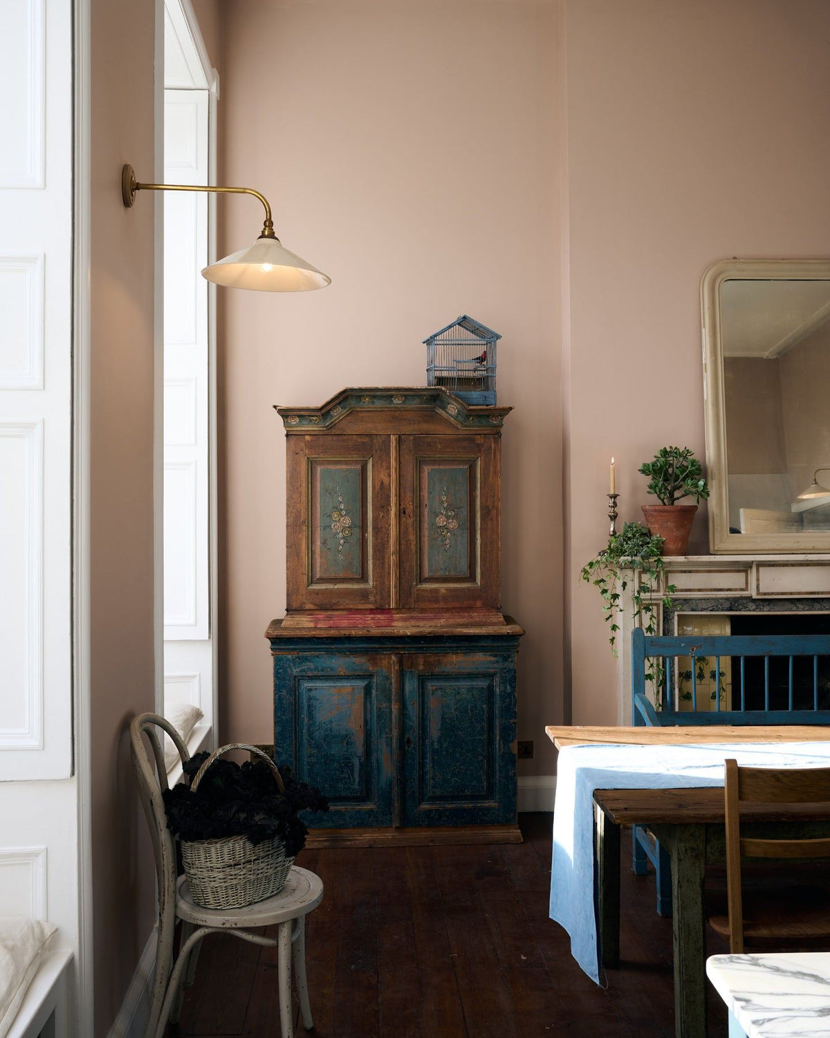 A grand room with high ceilings, soft pink walls, dark worn floorboards and antique furniture. A large Victorian-inspired wall light with a brass arm and a cream glazed ceramic shade is fixed to the wall by a tall window. TEST