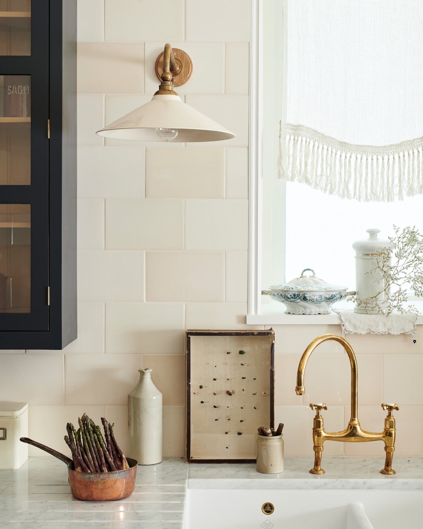 A straight-on view of a large Victorian-inspired wall light with a brass arm and a cream glazed ceramic shade fixed to a wall of cream subway tiles above a kitchen sink.