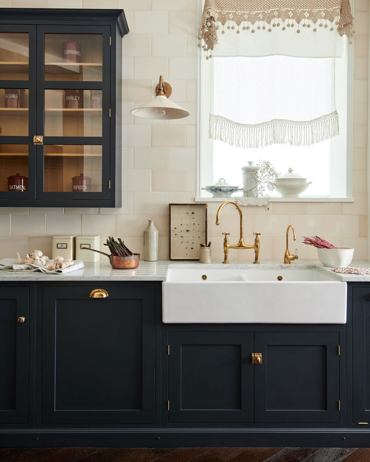 A straight-on view of a dark blue painted Shaker-style sink cupboard with a wide Belfast sink, an aged brass mixer tap, a wall of cream subway tiles and a large Victorian-inspired wall light with a brass arm and a cream glazed ceramic shade. TEST