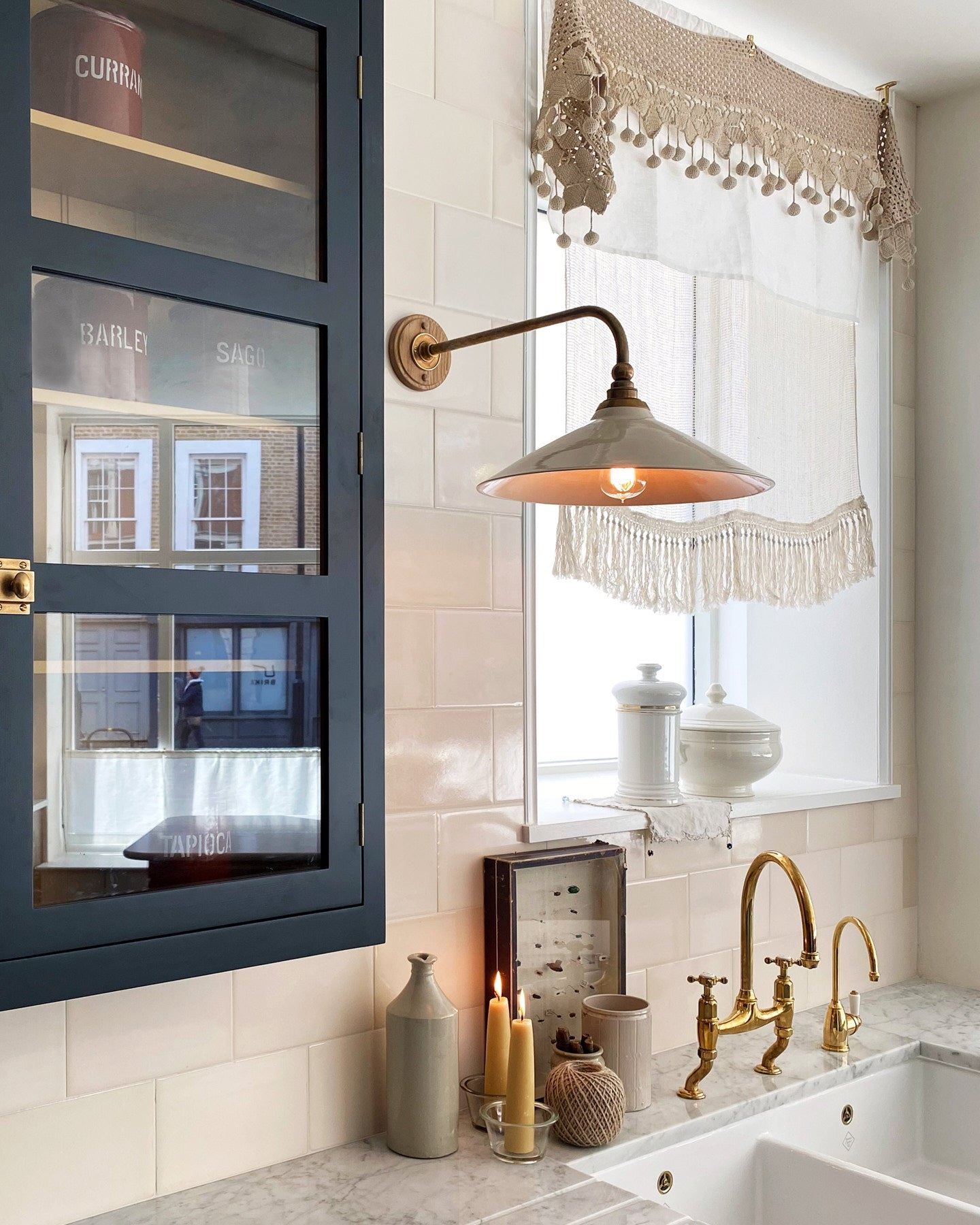 An oblique view of a large Victorian-inspired wall light with a brass arm and a cream glazed ceramic shade fixed to a wall of cream subway tiles above a kitchen sink.