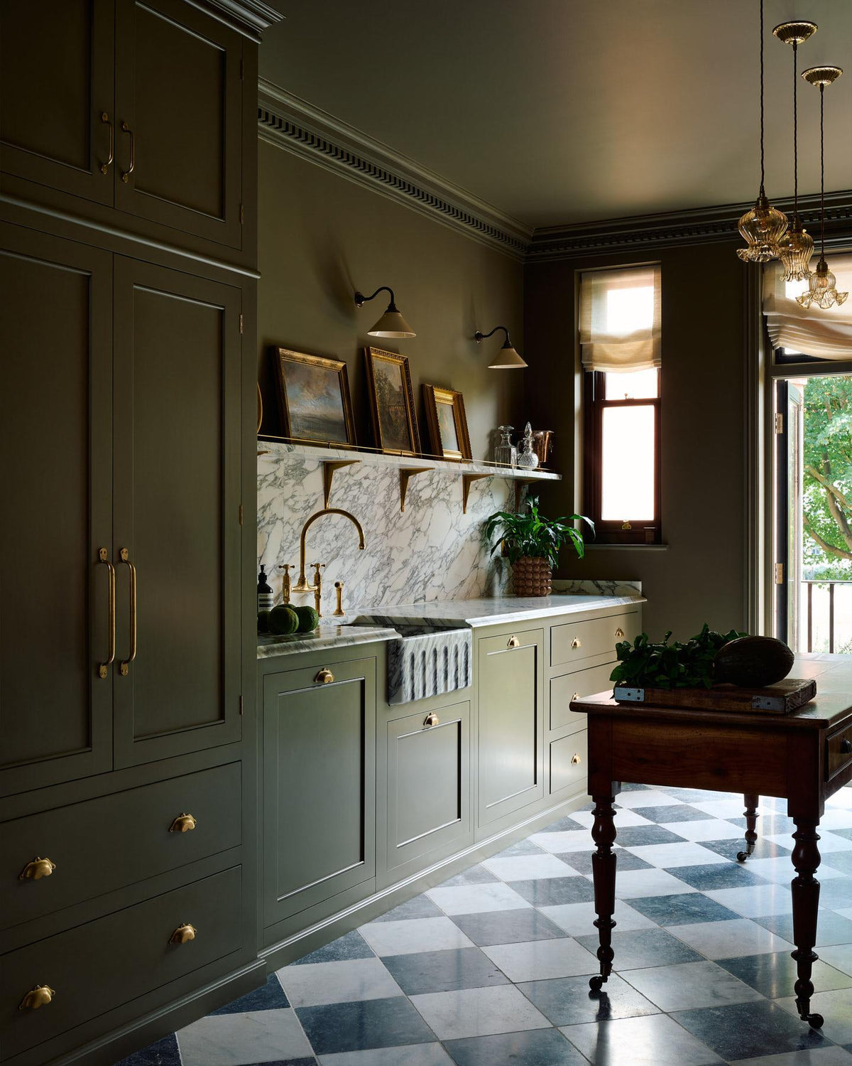 A straight-on view of a small wall light with an aged brass swan neck arm and a small cream glazed ceramic shade, fixed to a dark green wall above an Arabescato marble splashback and shelf, decorated with antique oil paintings in gilded frames. TEST