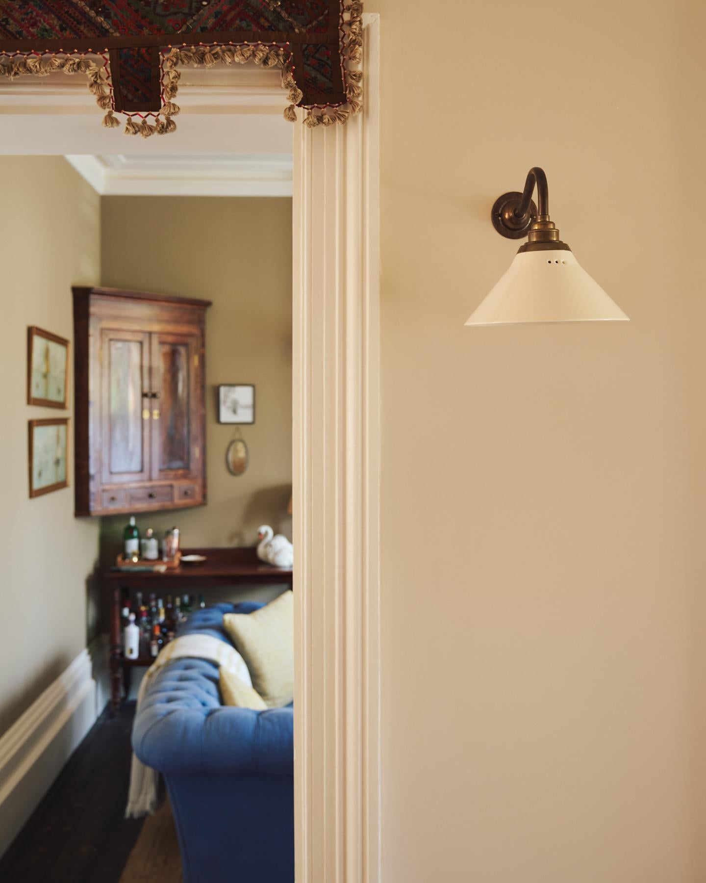 An oblique view of a small wall light with an aged brass swan neck arm and a small cream glazed ceramic shade, fixed to a cream wall.