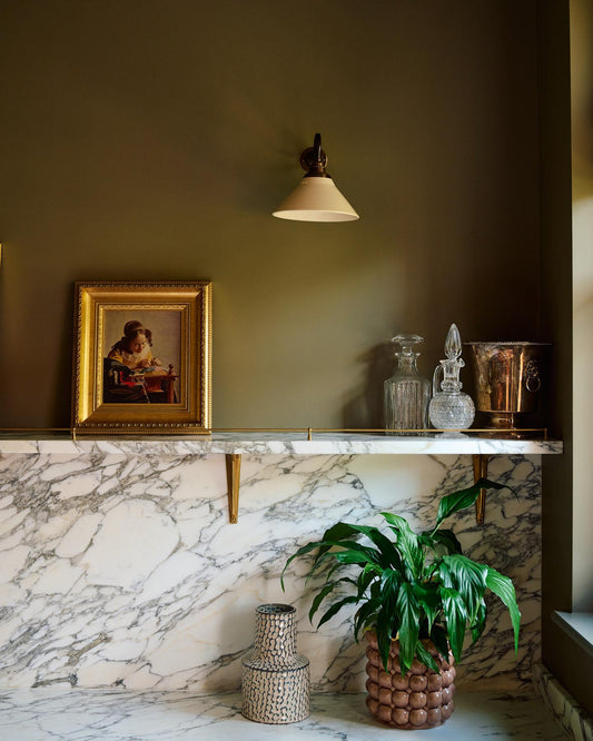 An oblique view of a small wall light with an aged brass swan neck arm and a small cream glazed ceramic shade, fixed to a dark green wall above an Arabescato marble shelf with glass bottles.