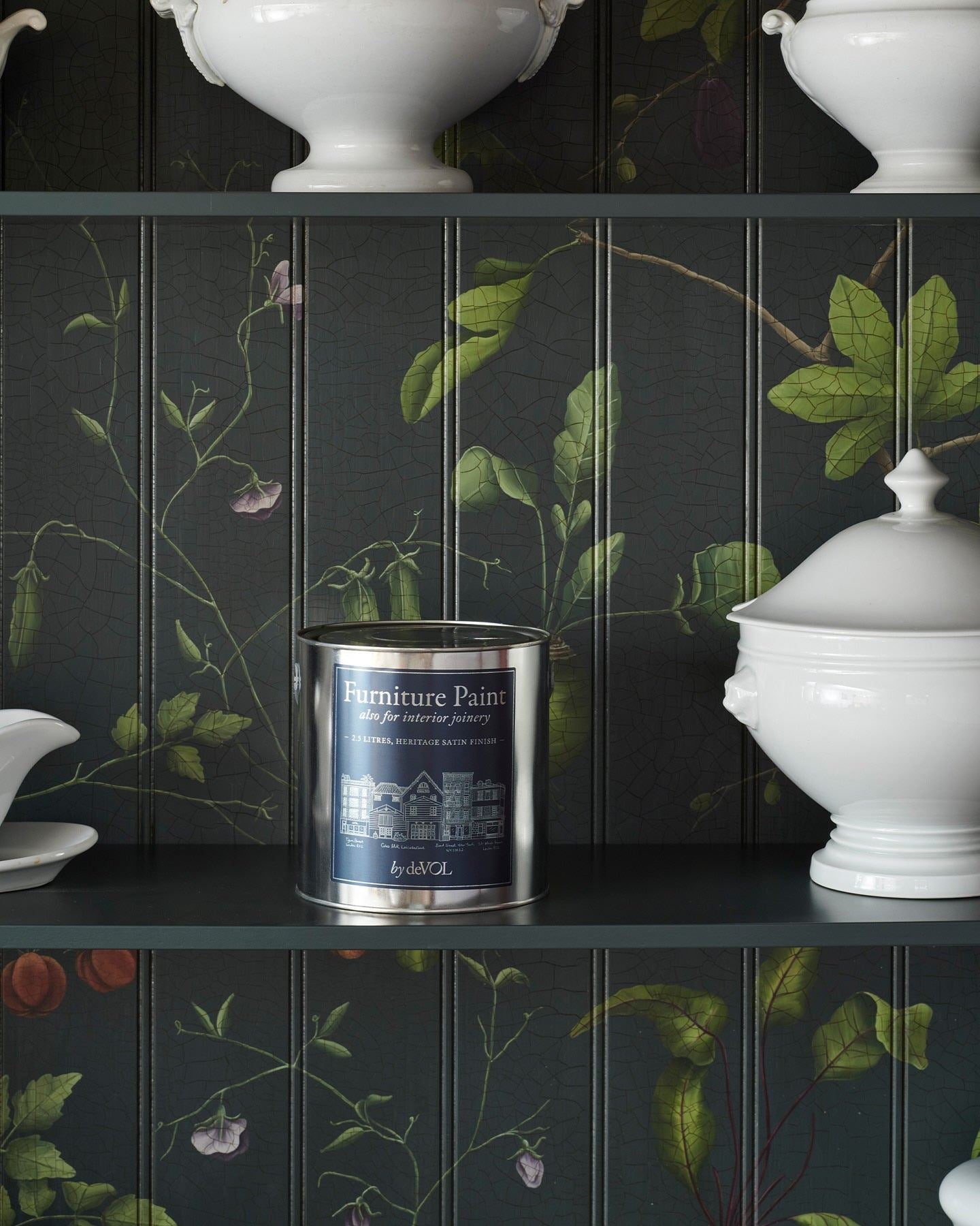 A silver paint tin with a rich blue label inside a dark green cupboard interior with hand-painted botanical flora on a tongue and groove-style backboard.