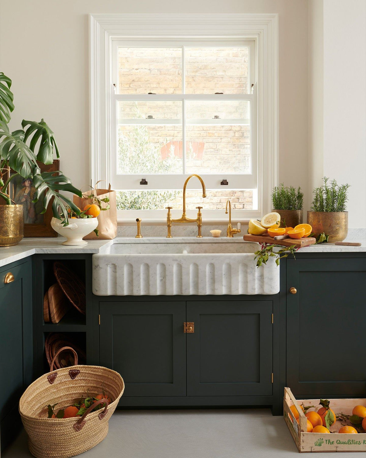 A dark green painted Shaker-style kitchen with light coloured walls, a cement floor, a wide Carrara marble sink with a fluted front, aged brass taps, a monstera plant and baskets of vibrant oranges. TEST
