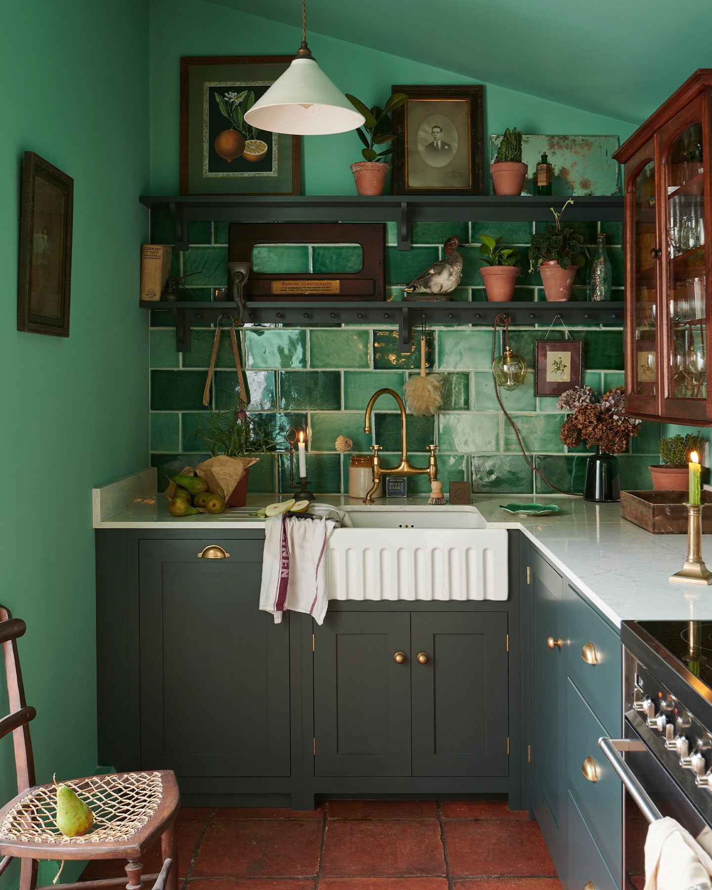 An eclectic cottage kitchen with dark green painted Shaker-style cupboards, arsenic green walls, glossy emerald green wall tiles, vintage bric-a-brac and a terracotta tiled floor.