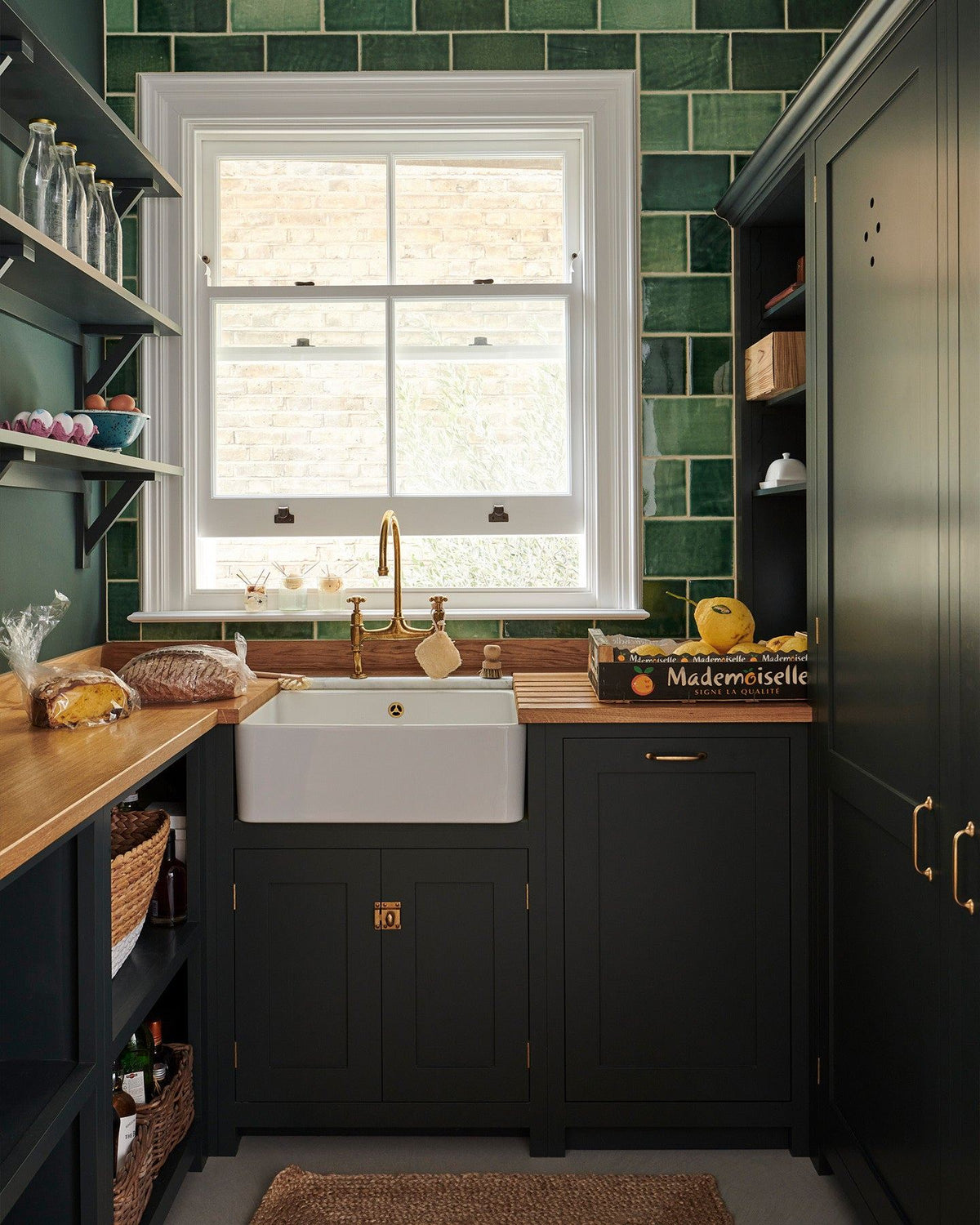 A moody utility kitchen with dark green painted Shaker-style cupboards, a small Belfast sink below a large sash window, hardwood worktops and a wall of glossy emerald green tiles. TEST