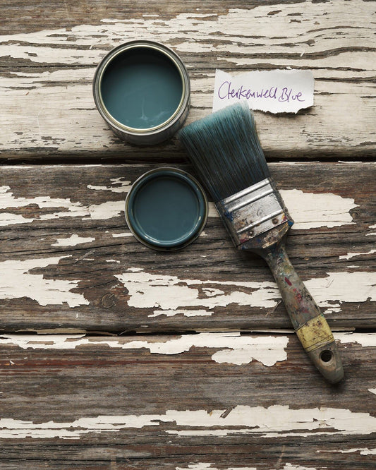 An overhead view of an open paint pot and a worn brush on a rustic tabletop. The green-tinged blue colour inside is labelled 'Clerkenwell Blue'.