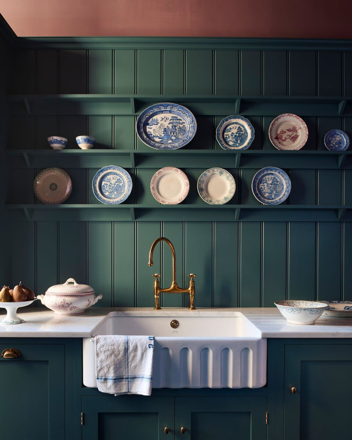 A straight-on view of a classically-styled kitchen sink with green-blue painted Shaker-style cupboards, tongue and groove wall panelling, shelving full of vintage crockery and wine red coloured walls. TEST
