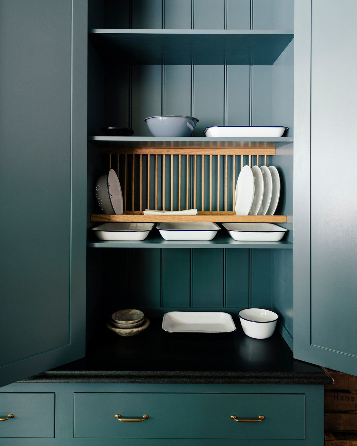 Looking into a Georgian-style pantry cupboard, painted inside and out in a green-tinged blue. Inside the cupboard there is wooden plate rack and a collection of white plates and enamel dishes. TEST