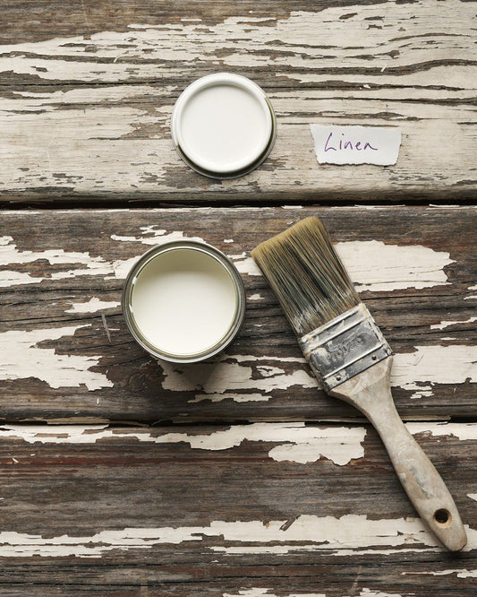 An overhead view of an open paint pot and a worn brush on a rustic tabletop. The light cream colour inside is labelled 'Linen'.