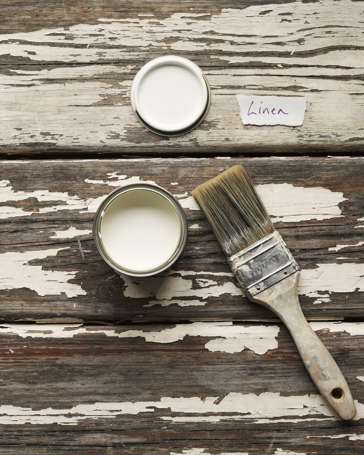 An overhead view of an open paint pot and a worn brush on a rustic tabletop. The light cream colour inside is labelled 'Linen'. TEST
