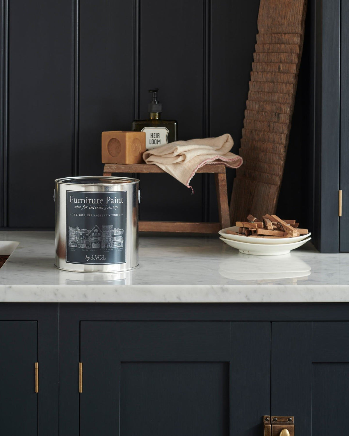 A close-up view of a silver paint tin with a blue label on a Carrara marble worktop, with dark blue painted cupboards and tongue and groove-style panelling behind. TEST