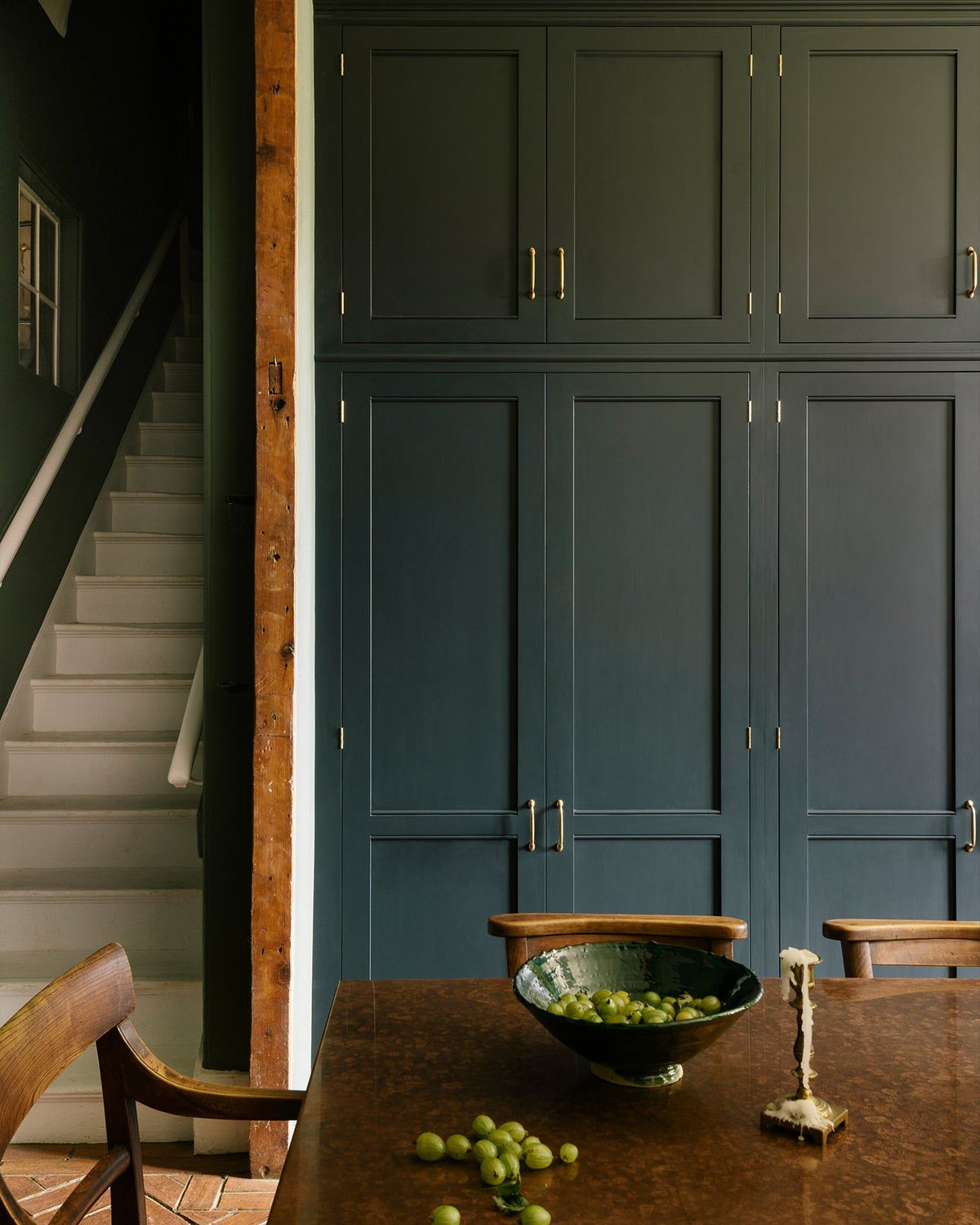 A straight-on view of tall Georgian-style kitchen pantry cupboards painted in a dark blue. There are white stairs to the left of the cupboard and an aged copper tabletop with a bowl of gooseberries in the foreground. TEST