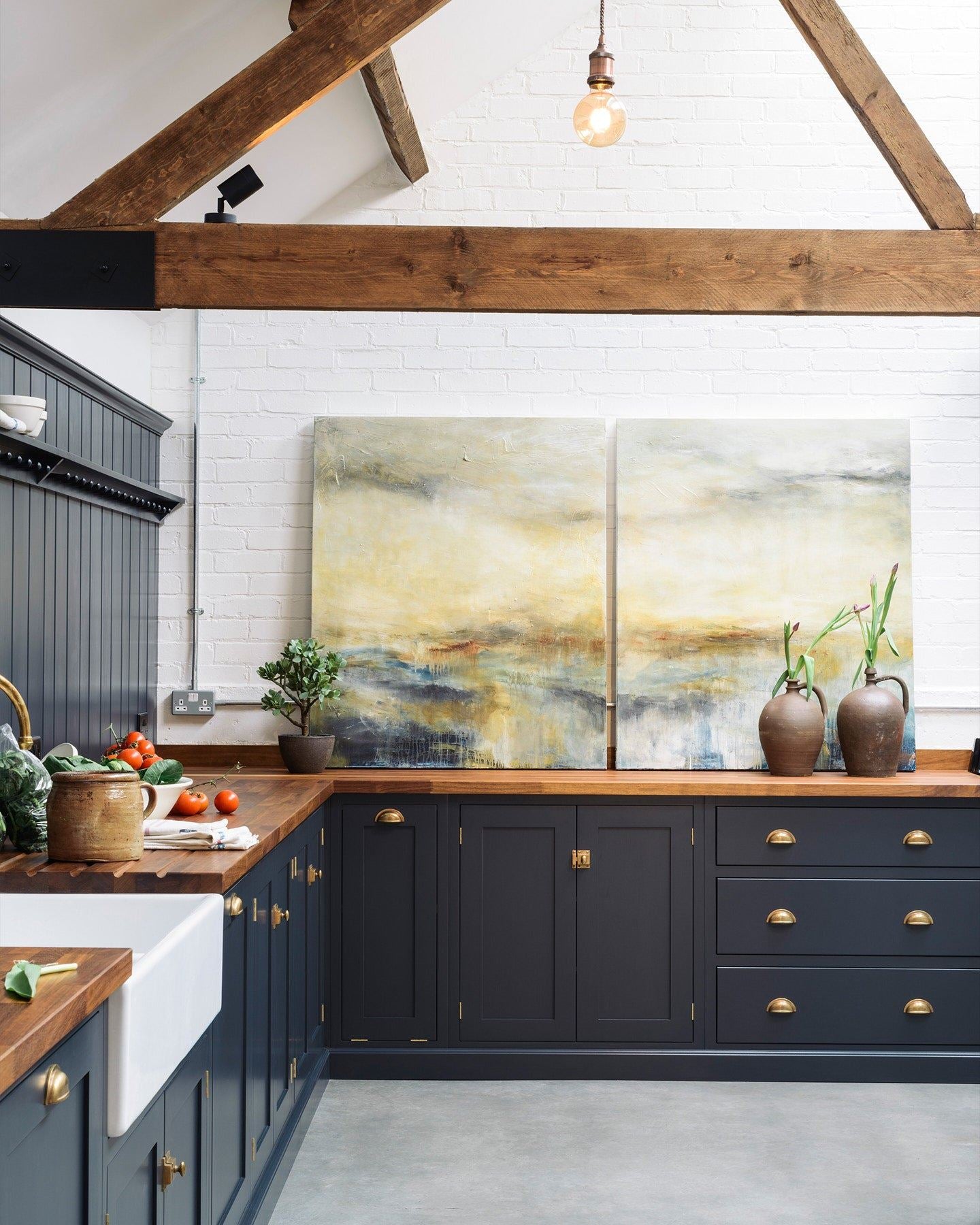 A large barn conversion with a simple Shaker-style kitchen, dark blue painted cupboards, brass hardware, rich hardwood worktops, white brick walls, a cement floor and old worn ceiling beams.