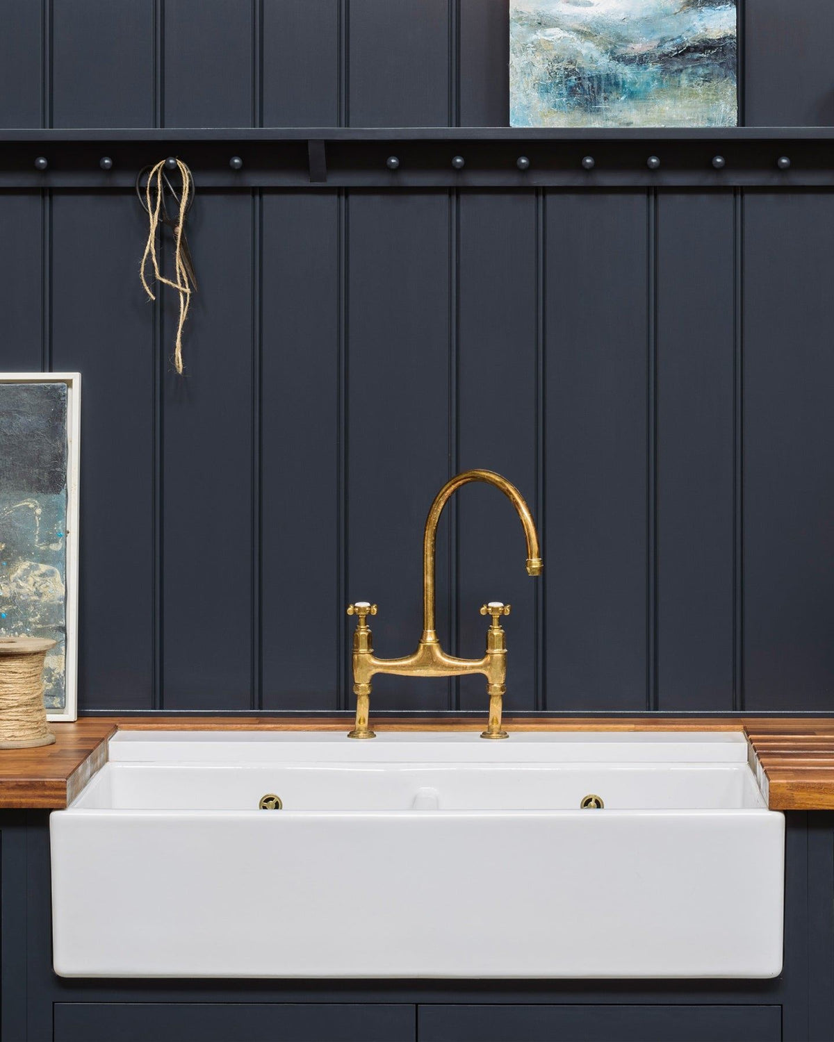 A close-up view of a wide ceramic Belfast sink with an aged brass tap, rich hardwood worktop and dark blue painted tongue and groove-style panelling behind. TEST