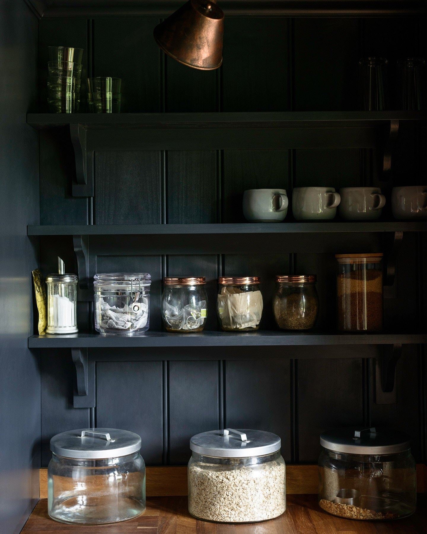 A dark blue painted tongue and groove panelled wall with Shaker-style shelving holding several jars of grains and cereals.