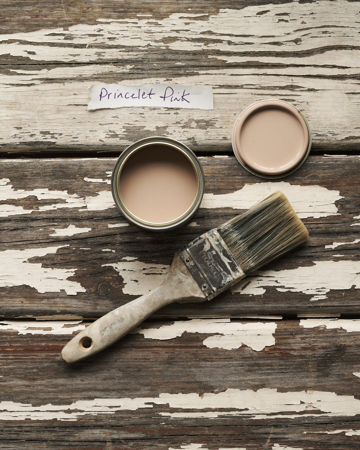 An overhead view of an open paint pot and a worn brush on a rustic tabletop. The soft pink colour inside is labelled 'Princelet Pink'. TEST