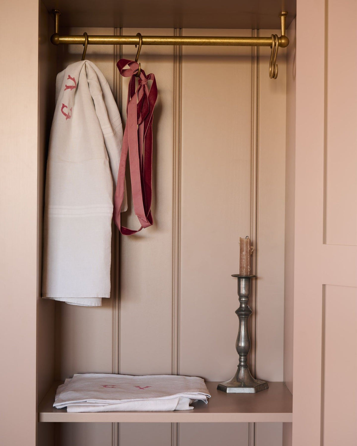 Looking into a Victorian-style pantry cupboard, painted inside and out in a soft pink colour. Inside the cupboard, there is a brass hanging rail with vintage linens and a bronze candlestick. TEST