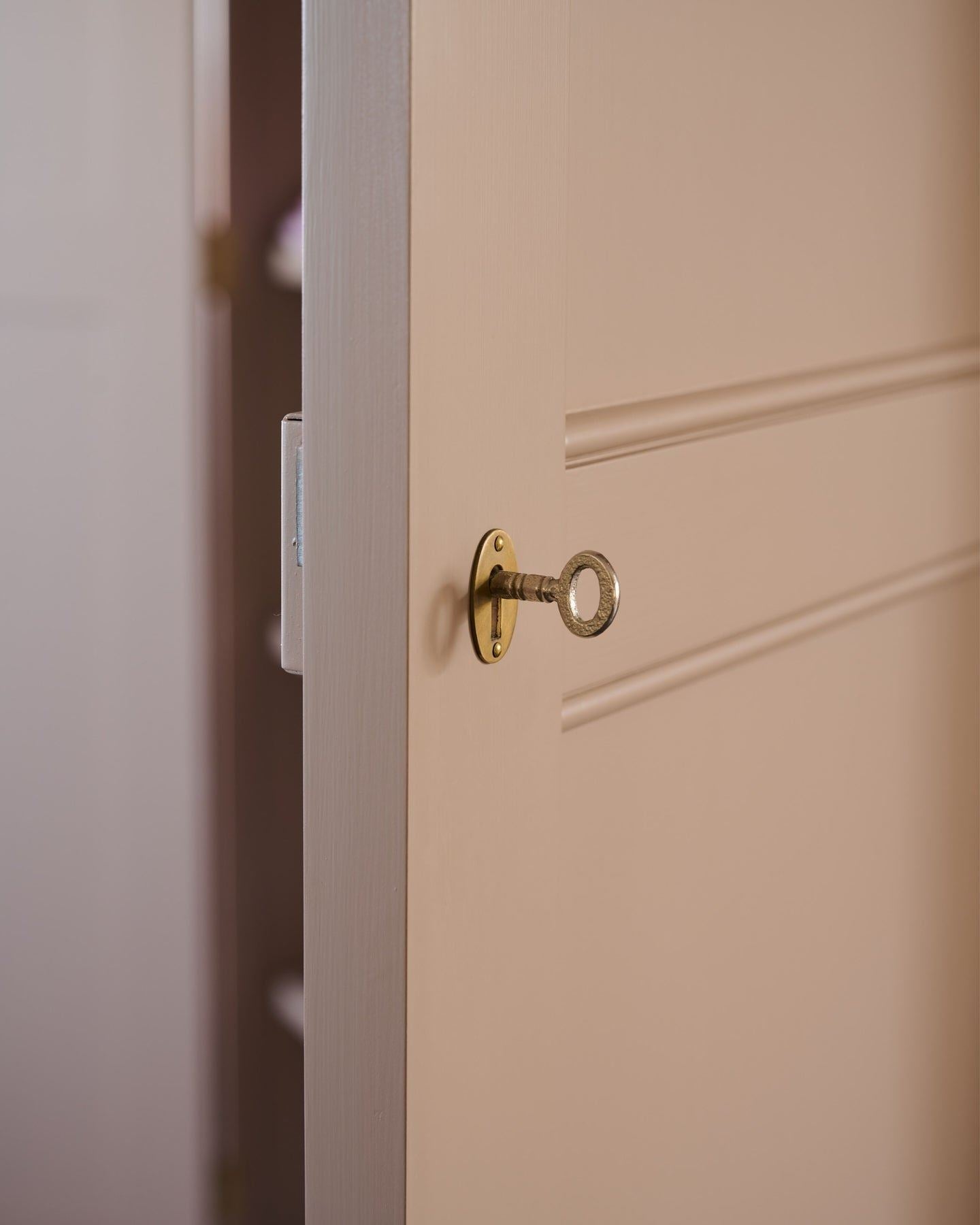 A close-up view of a soft pink coloured pantry door, ajar with a brass key inserted into an oval-shaped escutcheon.