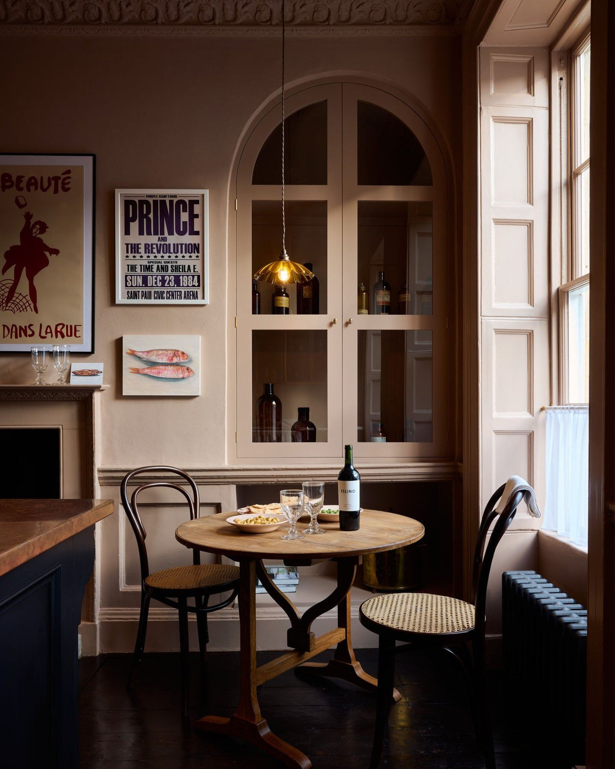 A straight-on view of a moody Georgian-style cafÈ-style corner with a small round pine dining table, pink walls and soft pink painted fitted glazed cupboard doors. TEST