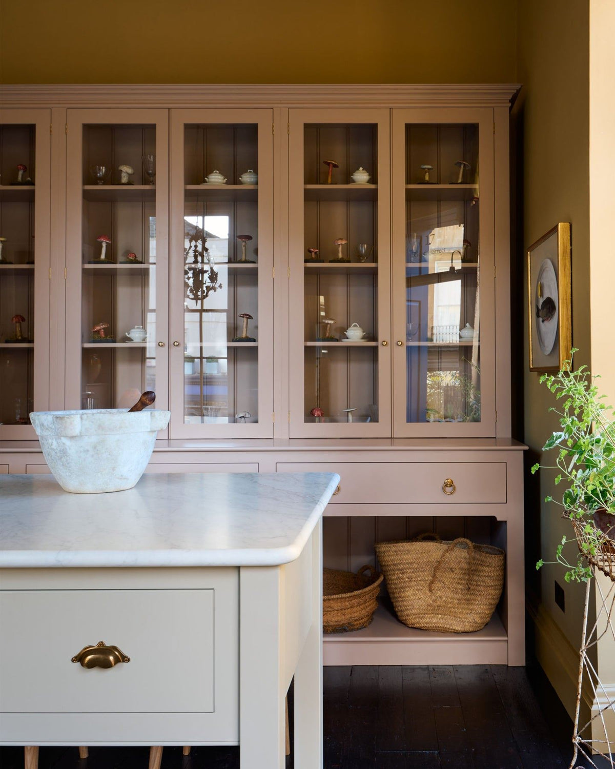 A straight-on view of a tall soft pink painted Georgian-style glazed dresser behind a large soft pink prep table with a Carrara marble worktop. TEST