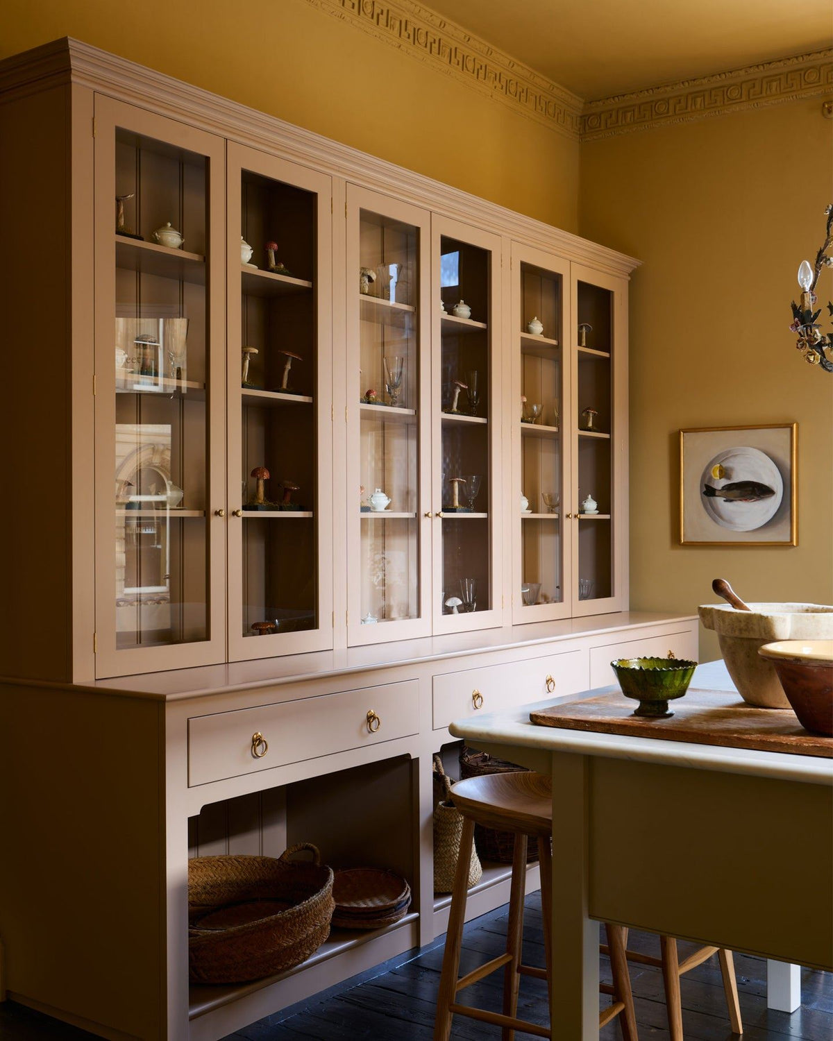 An oblique view of a tall soft pink painted Georgian-style glazed dresser in an atmospheric room with warm yellow walls. TEST