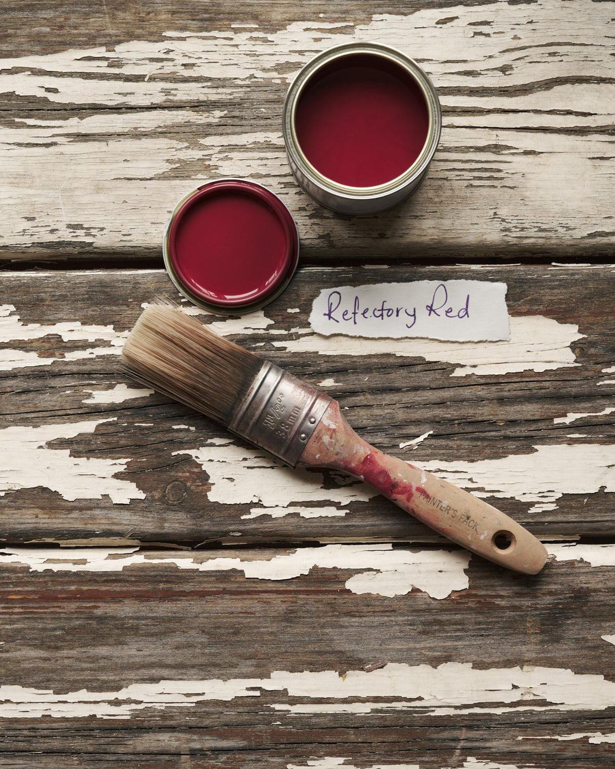 An overhead view of an open paint pot and a worn brush on a rustic tabletop. The rich red colour inside is labelled 'Refectory Red'. TEST