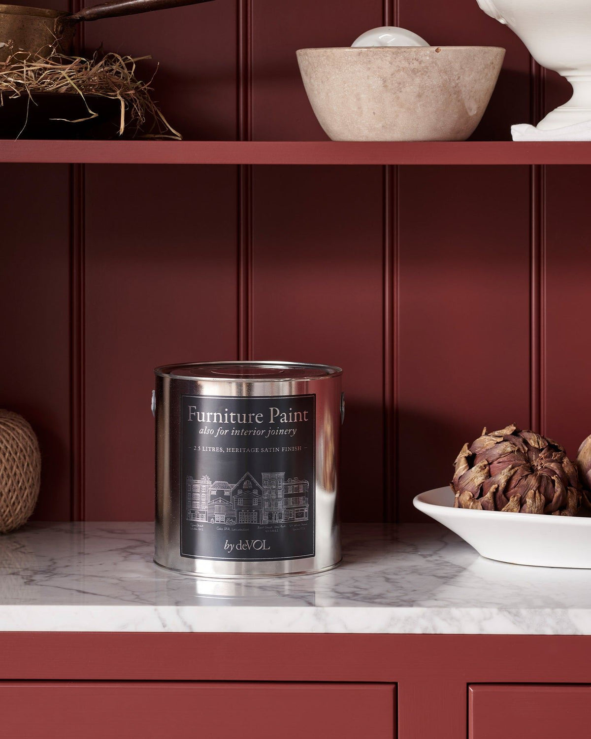 A close-up view of a silver paint tin with a blue label on a Carrara marble worktop, with rich red painted cupboards and tongue and groove-style panelling behind. TEST