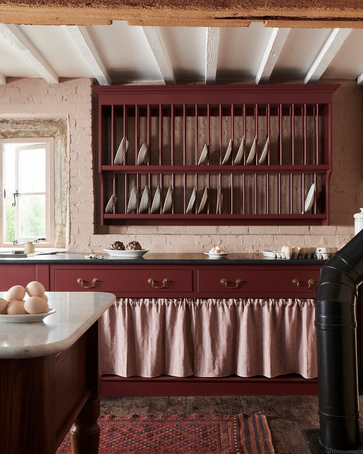 A straight-on view of a red painted Victorian-style kitchen with a traditional wall-mounted plate rack and wide drawers with open storage below. TEST