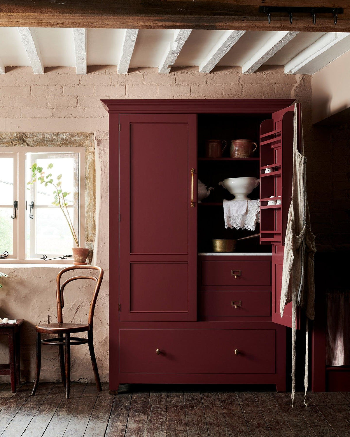 A red painted Georgian-style pantry cupboard with brass hardware and one door open, revealing white vintage crockery inside. TEST